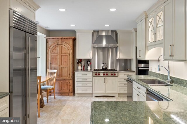kitchen with appliances with stainless steel finishes, wall chimney range hood, dark stone countertops, recessed lighting, and glass insert cabinets