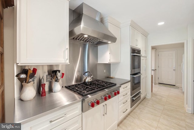 kitchen featuring wall chimney exhaust hood, stainless steel countertops, stainless steel appliances, a warming drawer, and white cabinetry