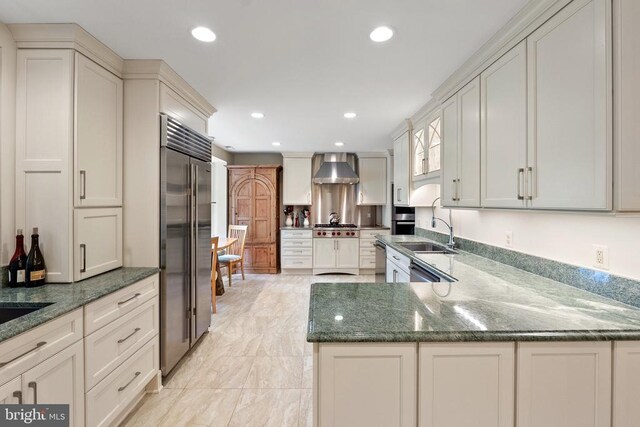 kitchen featuring appliances with stainless steel finishes, a sink, dark stone countertops, wall chimney range hood, and recessed lighting