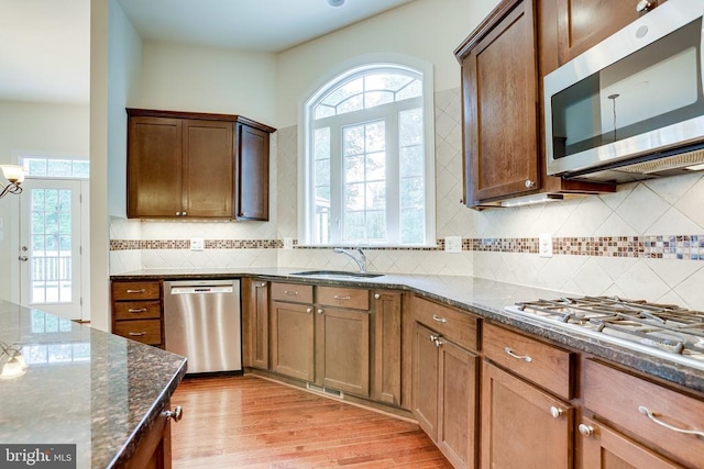 kitchen with appliances with stainless steel finishes, a sink, light wood finished floors, dark stone counters, and tasteful backsplash
