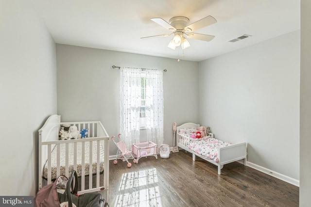 bedroom with a ceiling fan, baseboards, visible vents, wood finished floors, and a crib