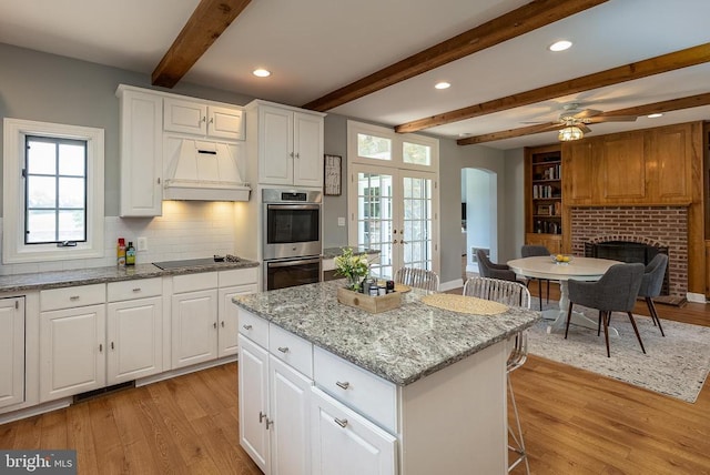kitchen featuring custom range hood, stainless steel double oven, arched walkways, beam ceiling, and light wood-style flooring