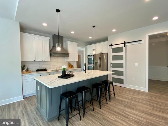 kitchen featuring wall chimney exhaust hood, appliances with stainless steel finishes, a barn door, light wood-style flooring, and recessed lighting