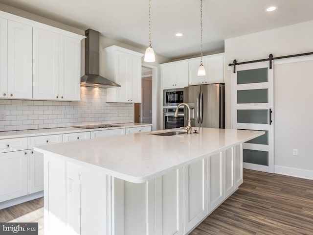 kitchen featuring wall chimney range hood, stainless steel appliances, a sink, a kitchen island with sink, and white cabinetry