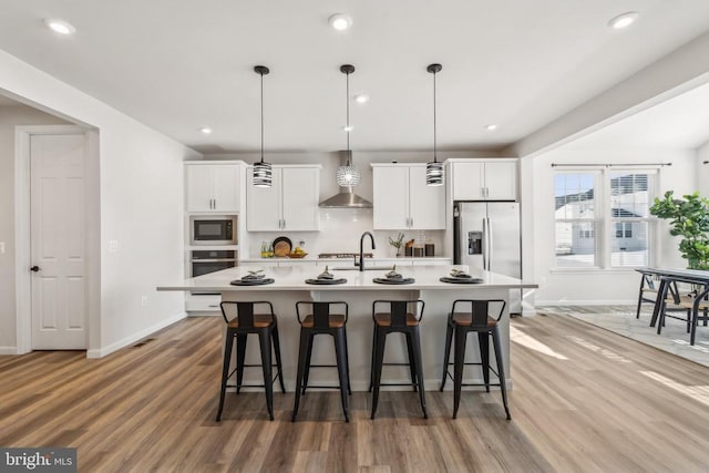 kitchen with stainless steel appliances, wall chimney exhaust hood, a kitchen island with sink, a breakfast bar area, and light wood-type flooring
