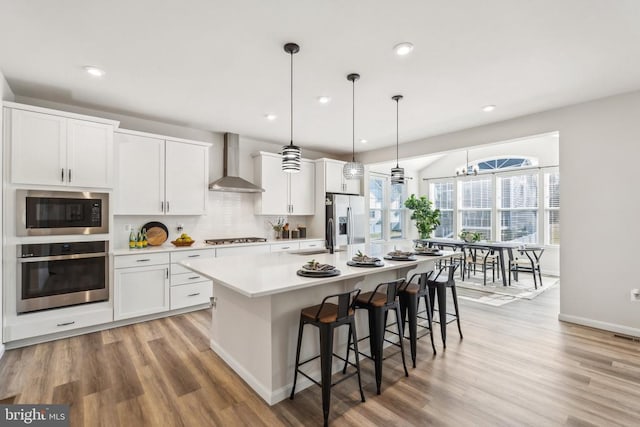 kitchen with stainless steel appliances, wall chimney exhaust hood, light wood finished floors, a kitchen island with sink, and recessed lighting