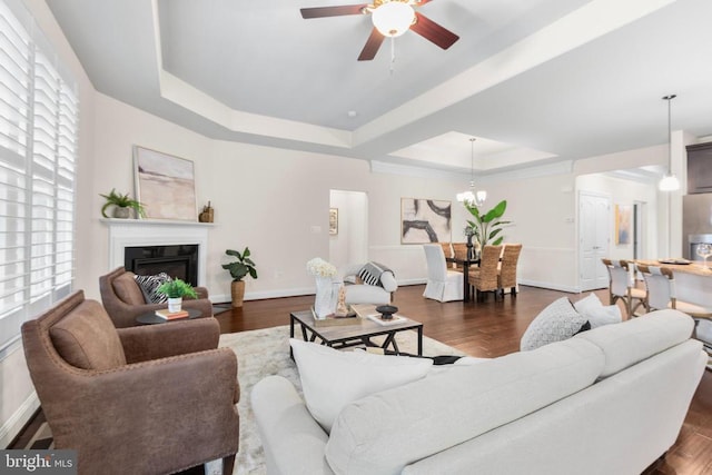 living area featuring baseboards, dark wood-type flooring, a tray ceiling, a fireplace, and ceiling fan with notable chandelier