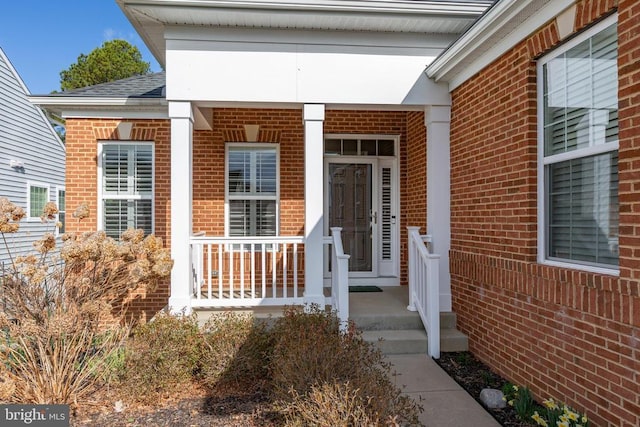 entrance to property featuring covered porch, roof with shingles, and brick siding