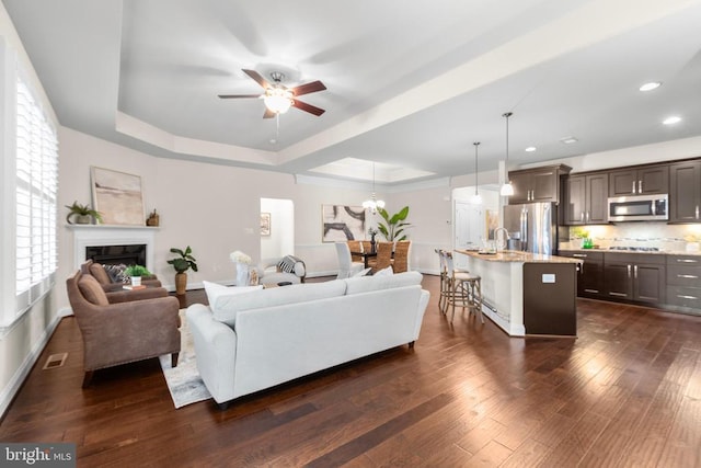 living room featuring a tray ceiling, a fireplace, recessed lighting, dark wood-type flooring, and ceiling fan with notable chandelier