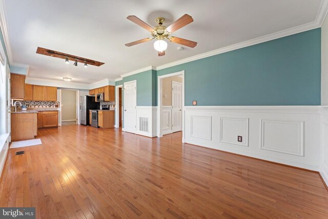 unfurnished living room featuring ceiling fan, light wood-type flooring, crown molding, a sink, and a wainscoted wall