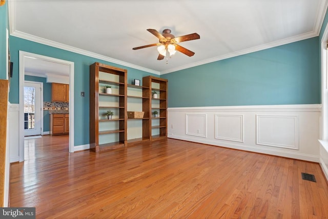 unfurnished room with ceiling fan, light wood-style flooring, wainscoting, and crown molding