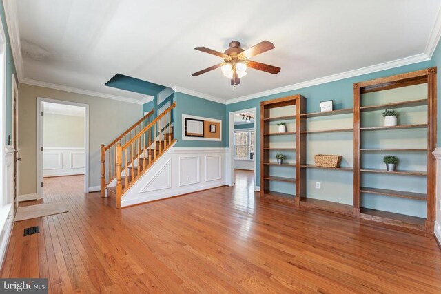 spare room featuring stairway, a ceiling fan, light wood-style flooring, crown molding, and a wainscoted wall