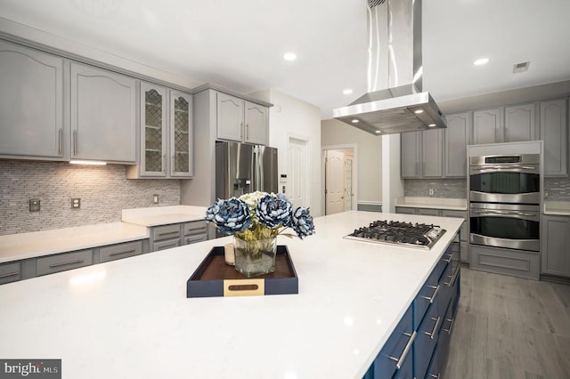 kitchen featuring appliances with stainless steel finishes, island exhaust hood, gray cabinets, decorative backsplash, and light countertops
