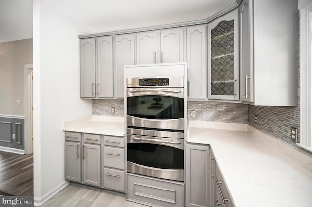 kitchen featuring gray cabinets, stainless steel double oven, decorative backsplash, light countertops, and glass insert cabinets