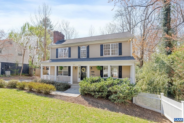 colonial-style house with covered porch, a chimney, fence, a front lawn, and central air condition unit