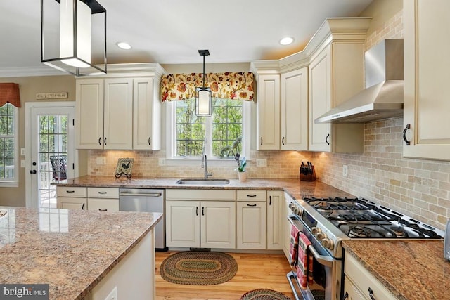 kitchen featuring appliances with stainless steel finishes, a sink, wall chimney exhaust hood, cream cabinets, and recessed lighting