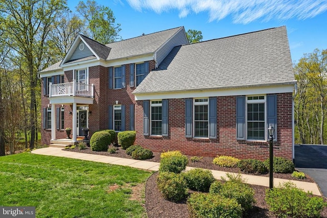 colonial house featuring a balcony, brick siding, a front yard, and roof with shingles