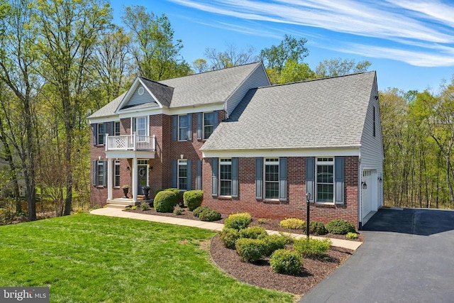 colonial home featuring a balcony, driveway, brick siding, a front lawn, and roof with shingles