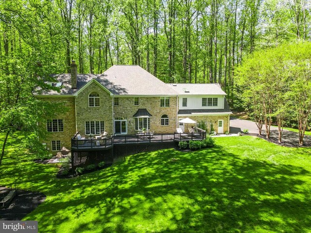  - back of house with a wooden deck, stone siding, a chimney, a lawn, and a forest view