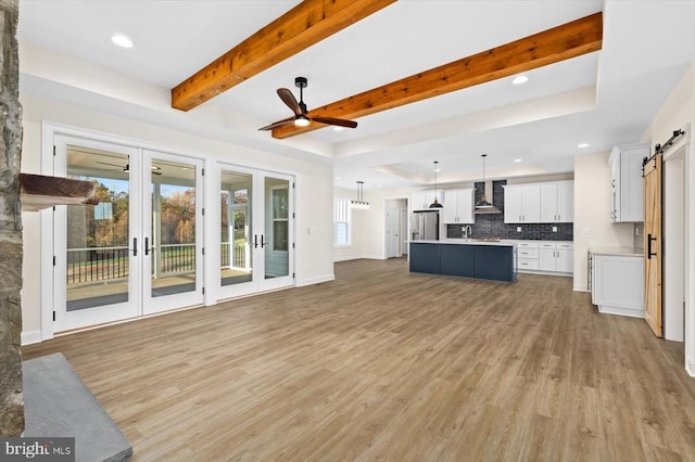 unfurnished living room with a barn door, light wood finished floors, french doors, ceiling fan, and beam ceiling