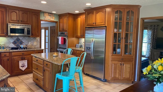  - kitchen featuring appliances with stainless steel finishes, light tile patterned flooring, light stone countertops, brown cabinets, and a textured ceiling
