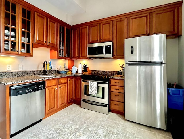 kitchen with glass insert cabinets, light tile patterned floors, a sink, dark stone countertops, and stainless steel appliances