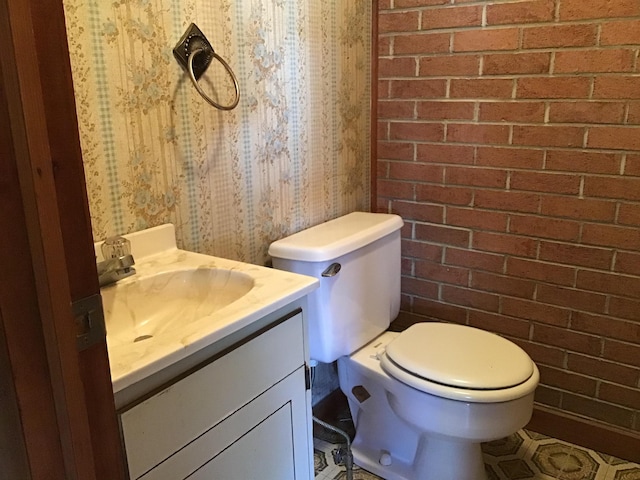bathroom with wallpapered walls, toilet, vanity, and brick wall