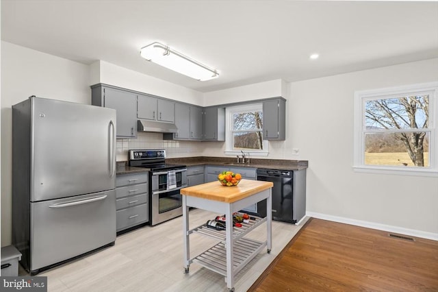 kitchen with gray cabinets, a sink, stainless steel appliances, under cabinet range hood, and dark countertops