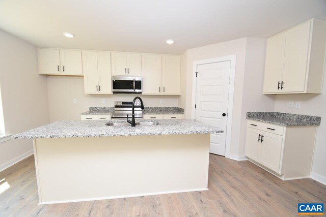kitchen featuring a sink, stainless steel appliances, light stone counters, an island with sink, and light wood-style floors
