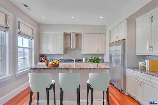 kitchen with a breakfast bar, stainless steel refrigerator with ice dispenser, visible vents, backsplash, and wall chimney range hood