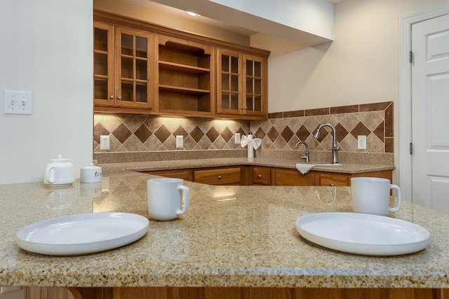 kitchen featuring decorative backsplash, glass insert cabinets, brown cabinets, and a sink