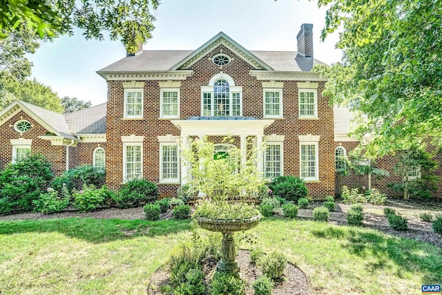 colonial inspired home featuring a front lawn, a shingled roof, brick siding, and a chimney