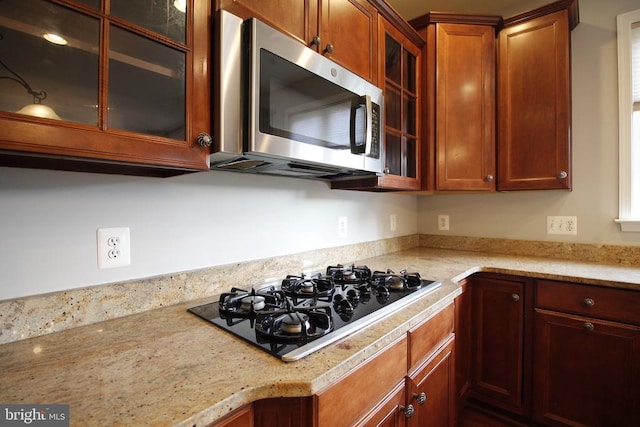  - kitchen with black gas stovetop, stainless steel microwave, glass insert cabinets, light stone countertops, and brown cabinets