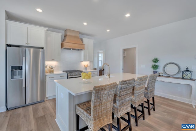 kitchen with premium range hood, light wood finished floors, stainless steel appliances, and a center island with sink