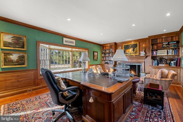  - office with wood finished floors, wainscoting, crown molding, a warm lit fireplace, and built in shelves