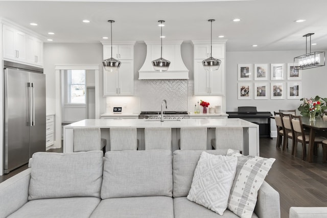 kitchen featuring built in fridge, custom range hood, decorative light fixtures, and a center island with sink