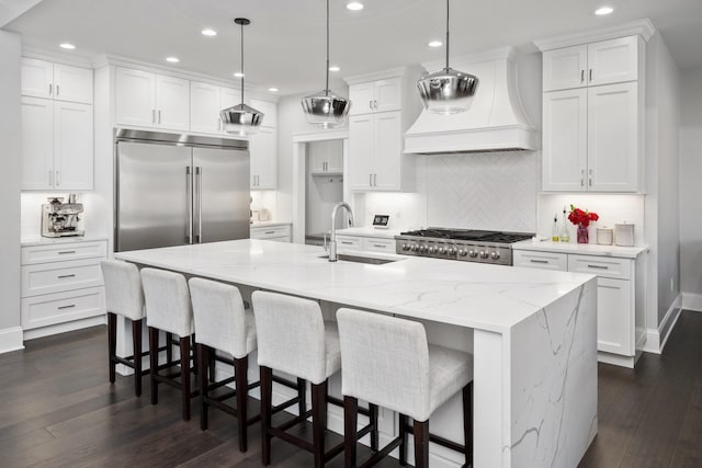 kitchen with built in fridge, decorative light fixtures, custom range hood, a sink, and a large island with sink