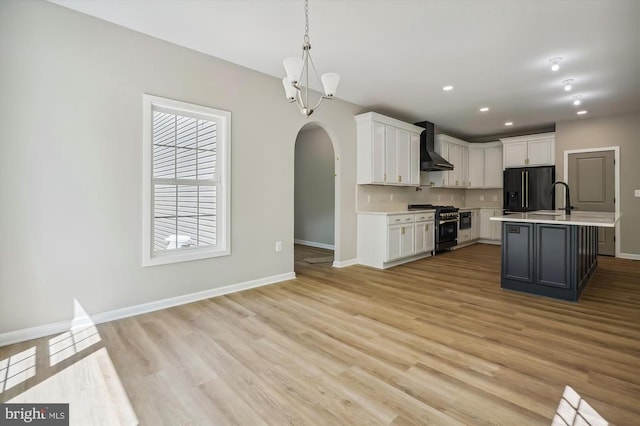  - kitchen featuring gas range, black refrigerator with ice dispenser, wall chimney range hood, arched walkways, and light countertops