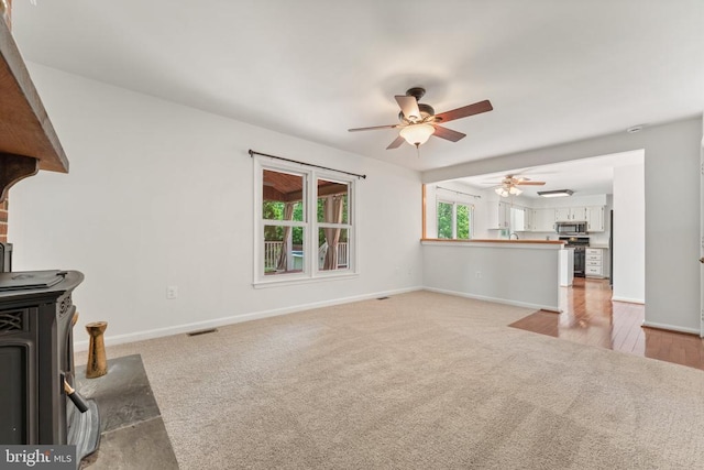 unfurnished living room with ceiling fan, light carpet, baseboards, and a wood stove