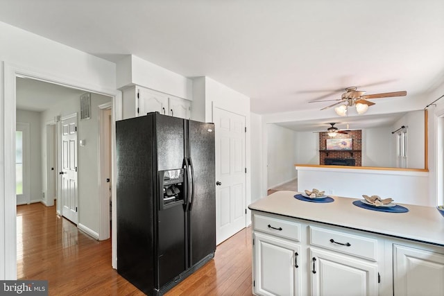 kitchen with black refrigerator with ice dispenser, a ceiling fan, a fireplace, white cabinets, and light wood-style flooring