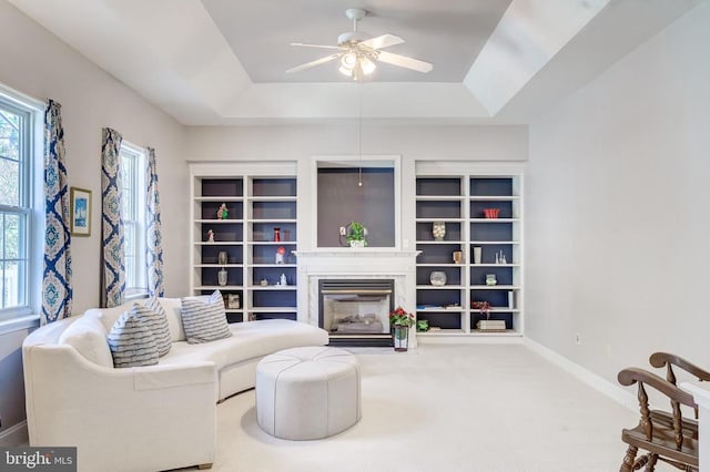 carpeted living area featuring a tray ceiling, a fireplace, a ceiling fan, baseboards, and built in features