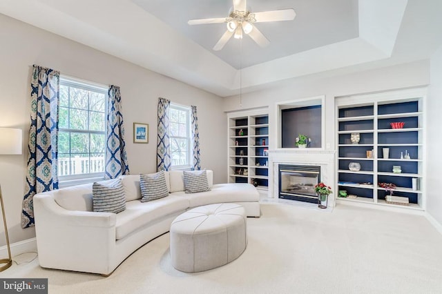 living room with a glass covered fireplace, a ceiling fan, carpet flooring, built in shelves, and a tray ceiling