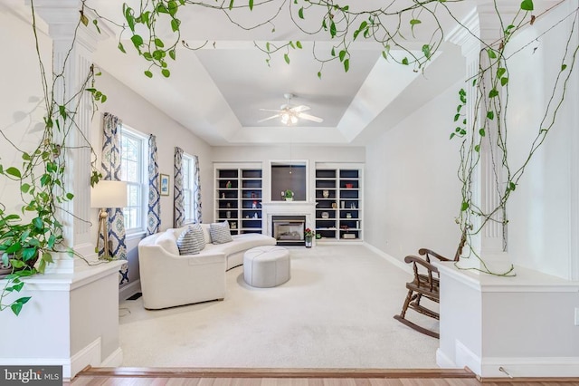 carpeted living area featuring a glass covered fireplace, baseboards, a ceiling fan, built in features, and a tray ceiling