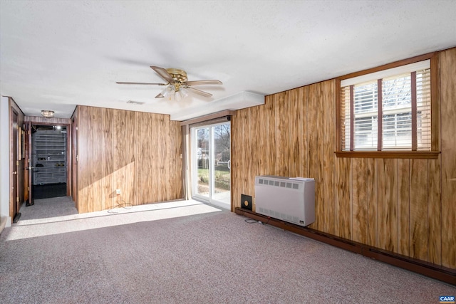 empty room featuring heating unit, carpet, a ceiling fan, wood walls, and visible vents