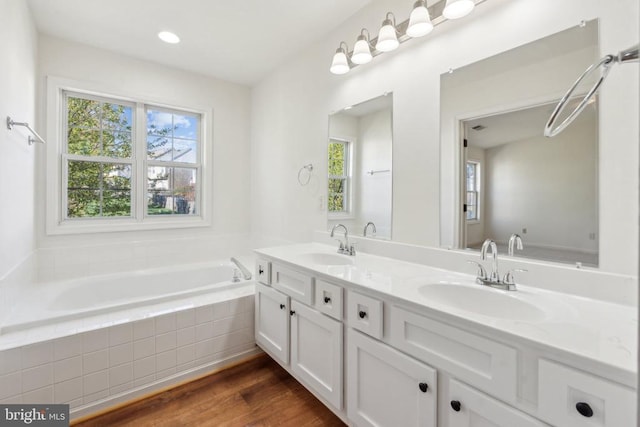 bathroom with plenty of natural light, a relaxing tiled tub, double vanity, wood finished floors, and recessed lighting