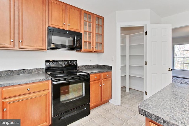 kitchen featuring light tile patterned floors, glass insert cabinets, dark countertops, brown cabinetry, and black appliances
