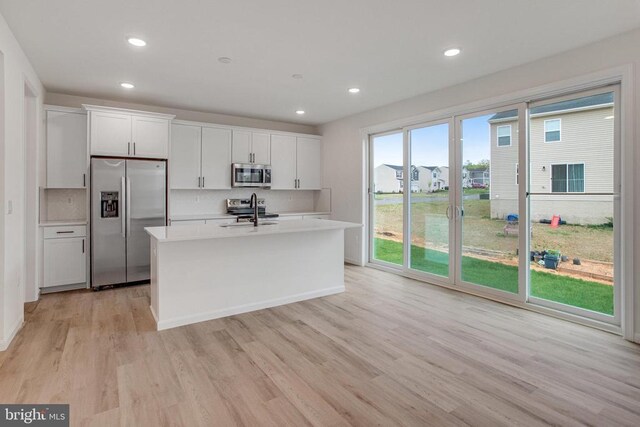 kitchen featuring stainless steel appliances, a kitchen island with sink, light countertops, light wood finished floors, and tasteful backsplash