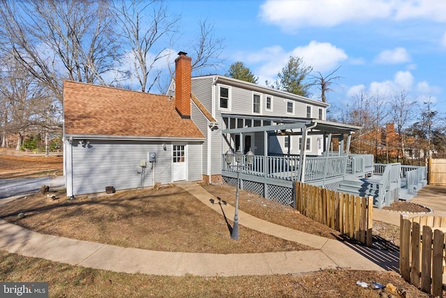 rear view of house with a shingled roof, a chimney, a deck, and fence