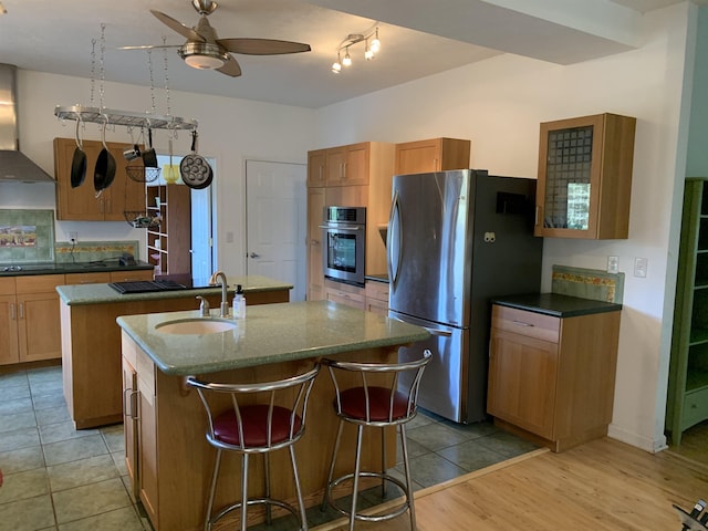 kitchen featuring wall chimney exhaust hood, sink, a center island with sink, ceiling fan, and stainless steel appliances