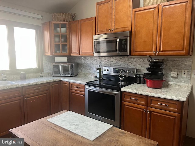 kitchen with backsplash, appliances with stainless steel finishes, brown cabinetry, a toaster, and glass insert cabinets